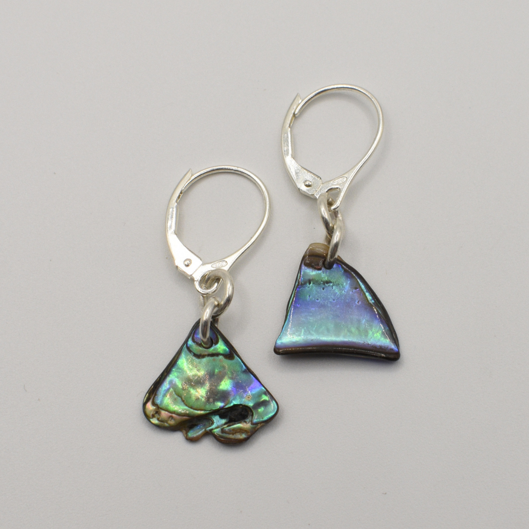 Salty Dreamers’ Earrings | Paua Shell Ngaru feature handmade silver hooks and unique New Zealand pāua shell charms—one irregular, one triangular—with iridescent green, blue, and purple on a white background.