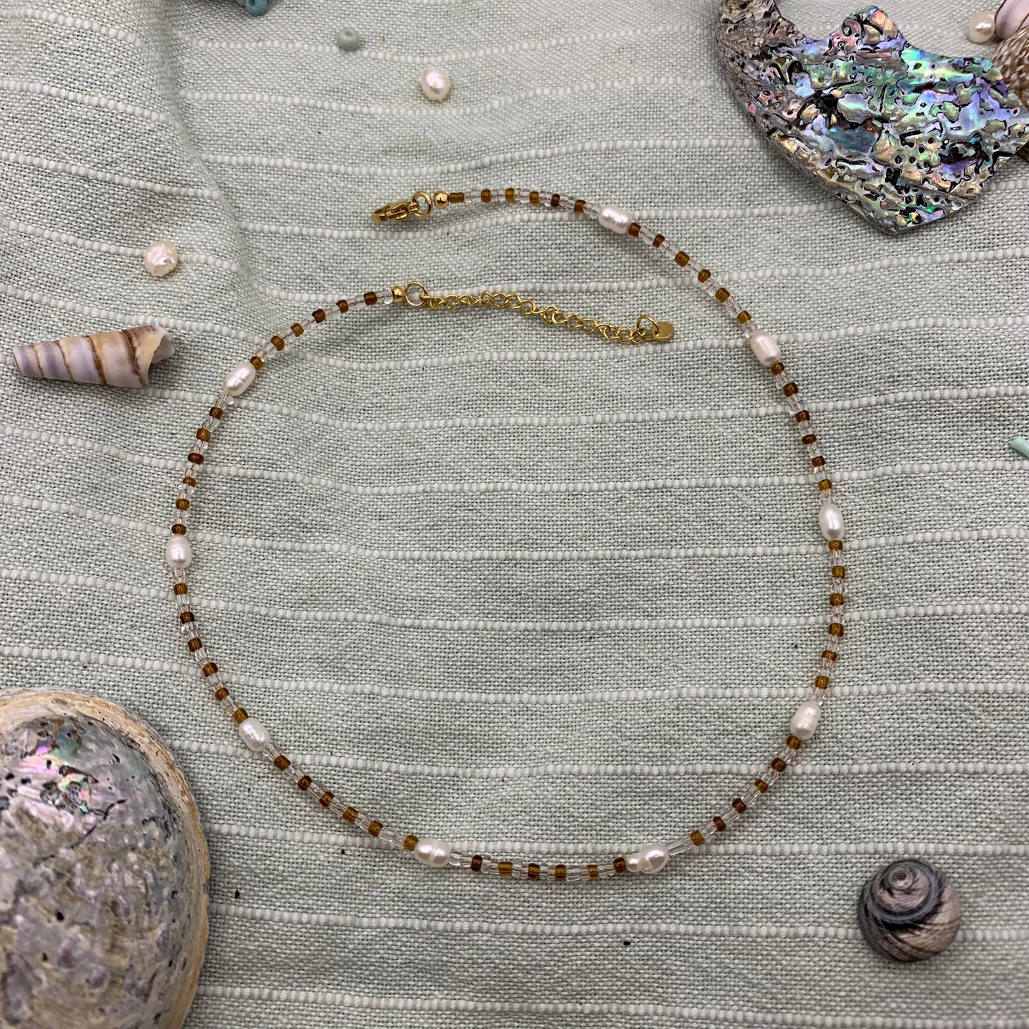 A handmade necklace with a combination of glass beads and freshwater pearls, featuring a golden stainless steel chain, displayed against a textured green background with seashells and other small items.