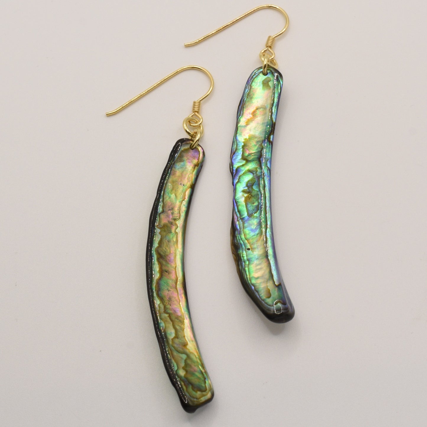 Salty Dreamers Earrings | Paua Shell Taurami feature long, curved handmade pāua shell with green, blue, and gold iridescence from NZ abalone, hanging from gold hooks and displayed on a white background.