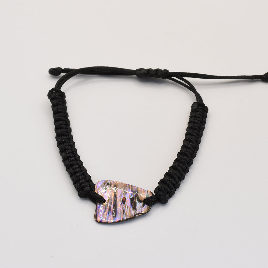 The Salty Dreamers Bracelet | Paua Shell Toa is a handmade black braided cord bracelet featuring an iridescent, irregular New Zealand paua shell centerpiece, shown on a plain white background.