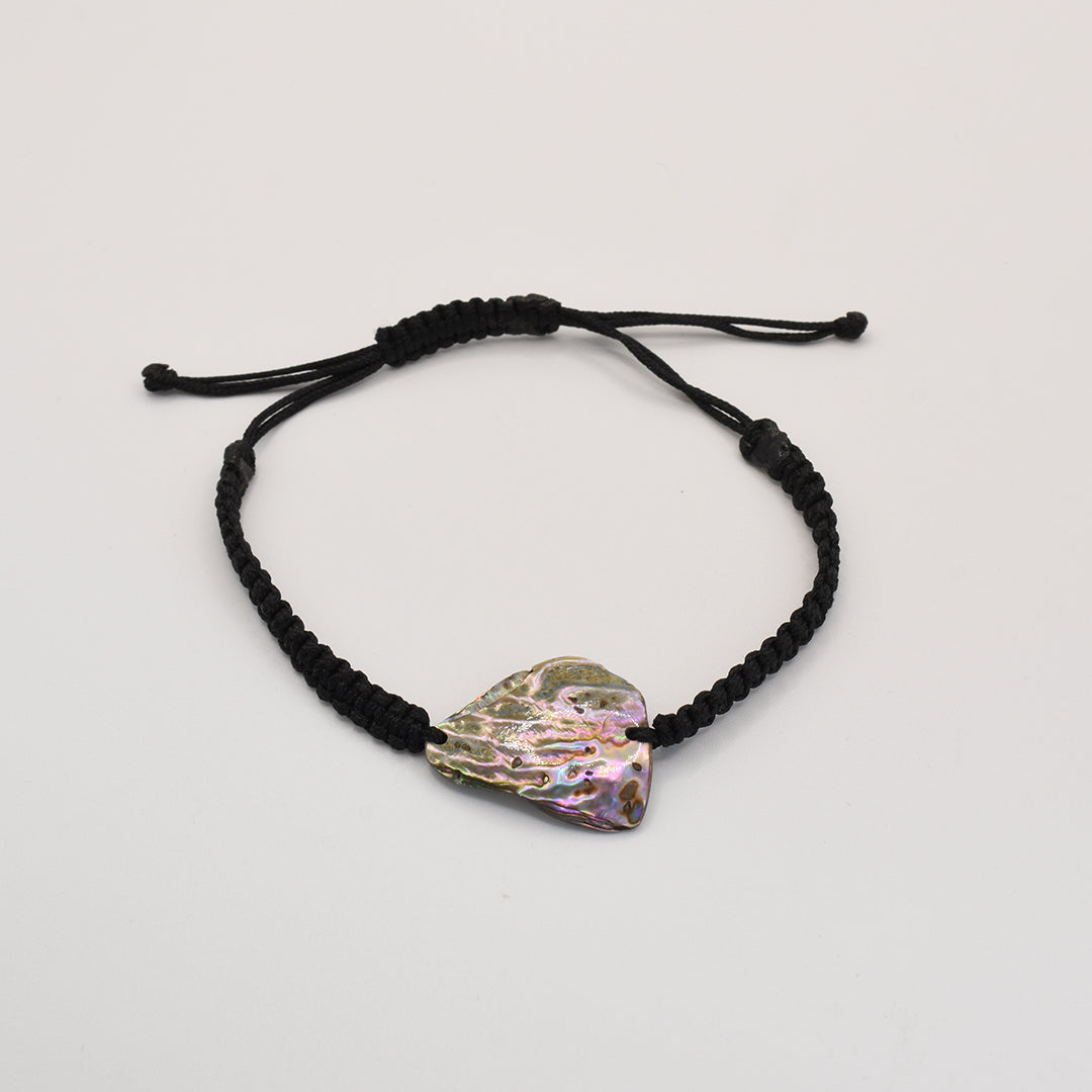 The Salty Dreamers Bracelet | Paua Shell Kaha features an irregular, iridescent pāua shell on a black braided cord, reflecting the beauty of New Zealand jewelry against a plain white background.