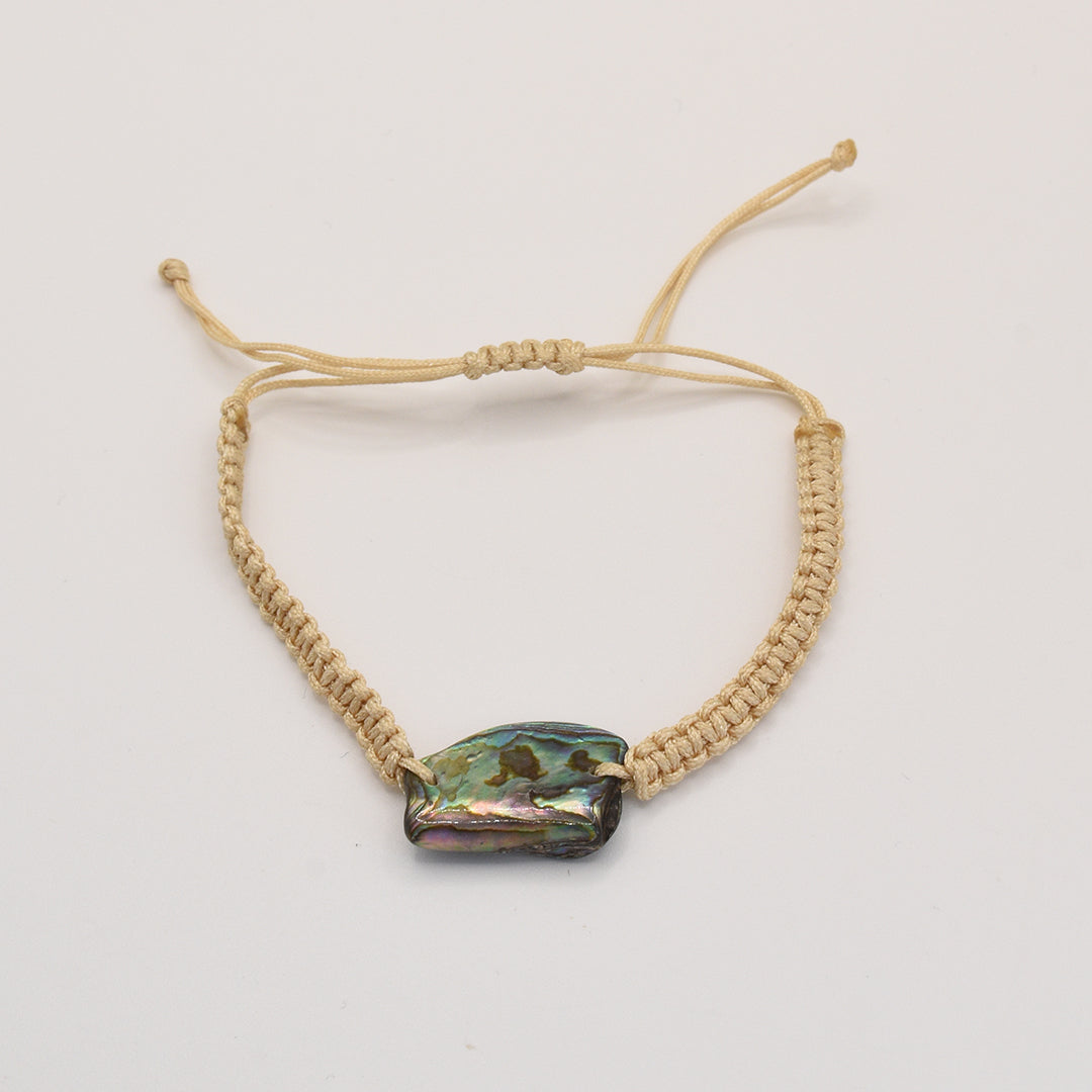 The Bracelet | Paua Shell Rangi by Salty Dreamers is a handmade beige macrame bracelet with a polished, irregular pāua shell bead at the center, shown on a plain white background.