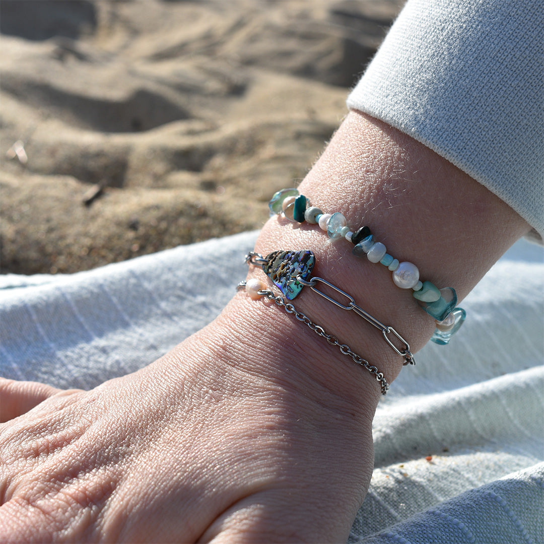 A close-up of a wrist wearing the Salty Dreamers Bracelet | Ocean Bamboo Blue, featuring handmade beads and a silver heart charm, resting on light fabric near sandy ground.