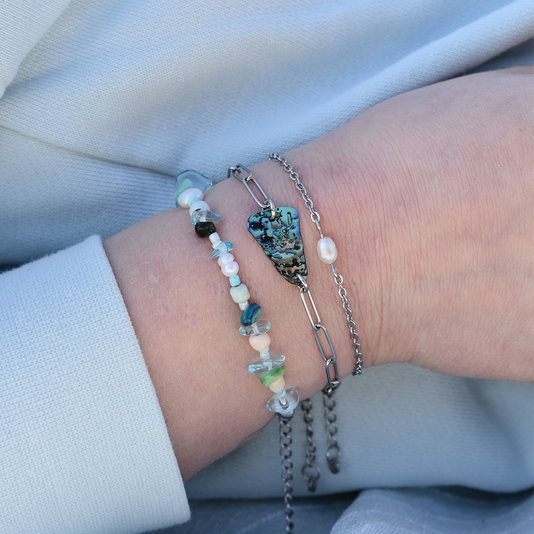A close-up of a wrist wearing Salty Dreamers Bracelet | Ocean Bamboo Blue alongside beaded and pearl bracelets, plus an abalone shell chain. The person is dressed in light blue fabric.