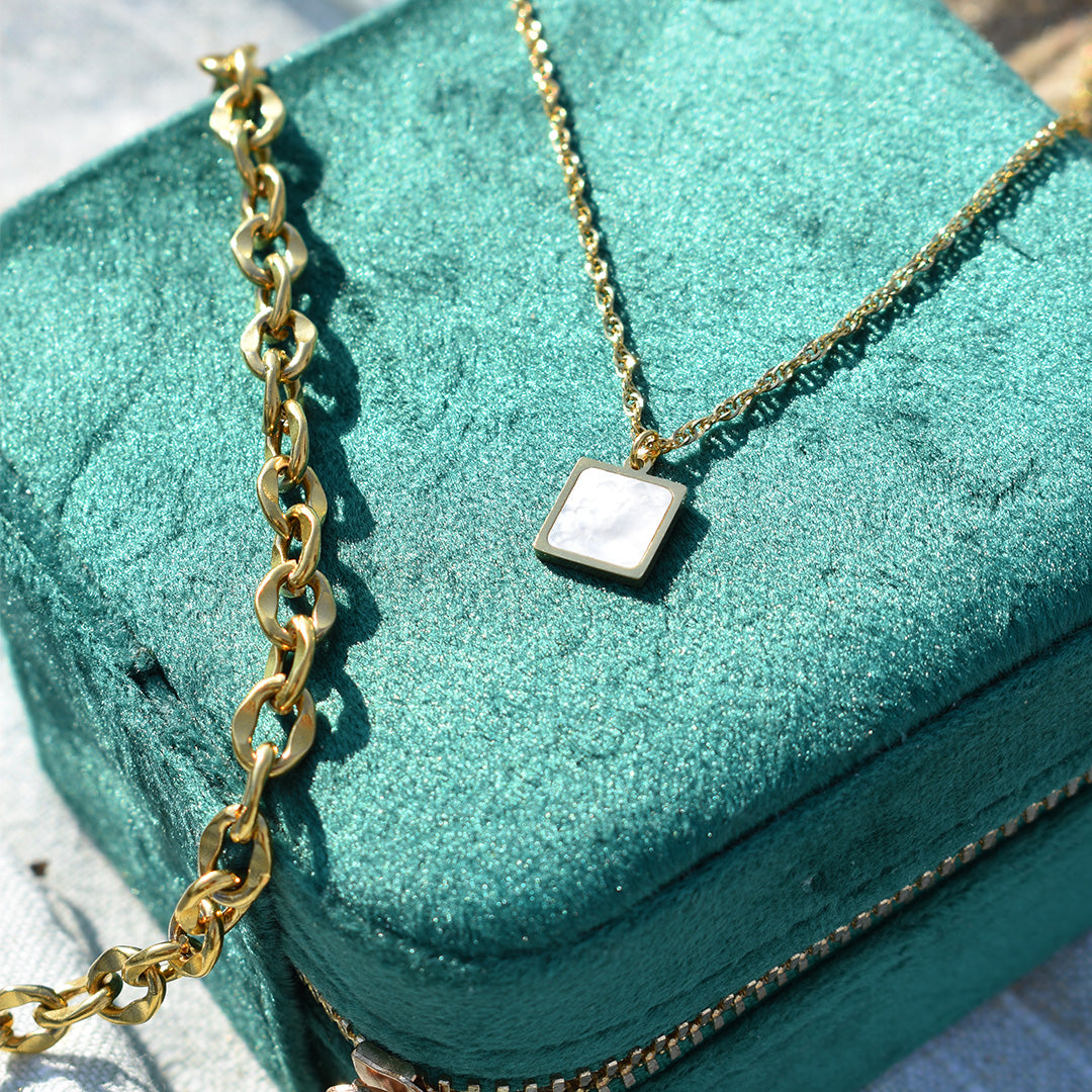 A gold chain necklace with a square mother-of-pearl pendant rests on a textured green jewelry box, next to Salty Dreamers Bracelet | Stainless Waves.