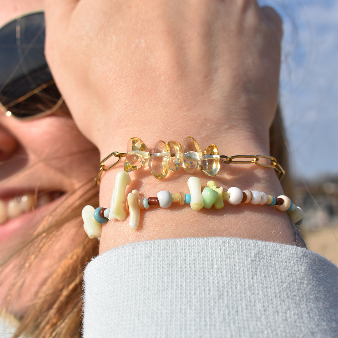A close-up of a smiling person outdoors wearing Salty Dreamers’ Bracelet | Ocean Bamboo Rainbow—two colorful handmade bead bracelets, featuring transparent yellow and pastel-white beads, sparkling on their wrist.