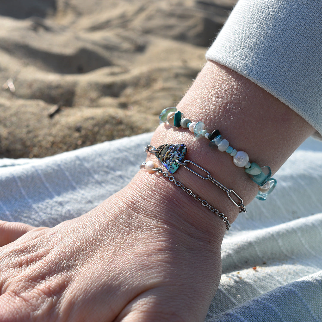 A close-up of a wrist wearing the Salty Dreamers Bracelet | Cutie Pearly Silver, featuring ocean-inspired details, resting on striped cloth at the beach.