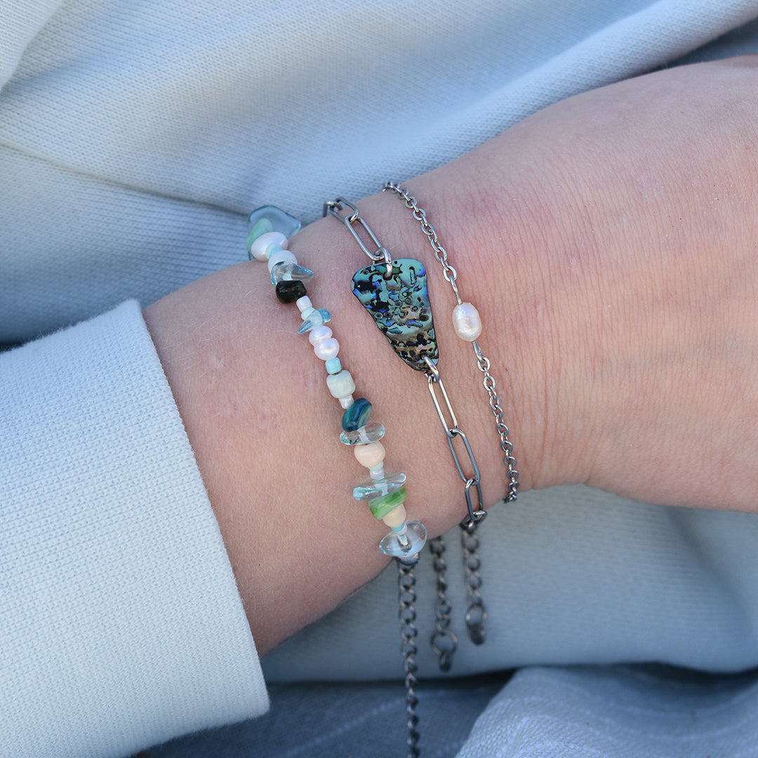 A close-up of a wrist wearing Salty Dreamers Bracelet | Cutie Pearly Silver, a handmade pearl and abalone shell design with mixed beads, an abalone heart charm, and a delicate chain. Light blue fabric serves as the backdrop.