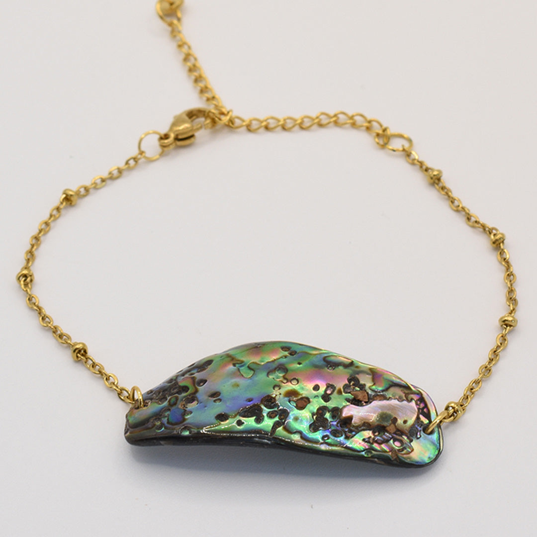 The Paua Shell Mahaki bracelet by Salty Dreamers features an iridescent pāua shell centerpiece in green, blue, and purple on a gold chain with a lobster clasp—a striking piece of handmade New Zealand jewelry.