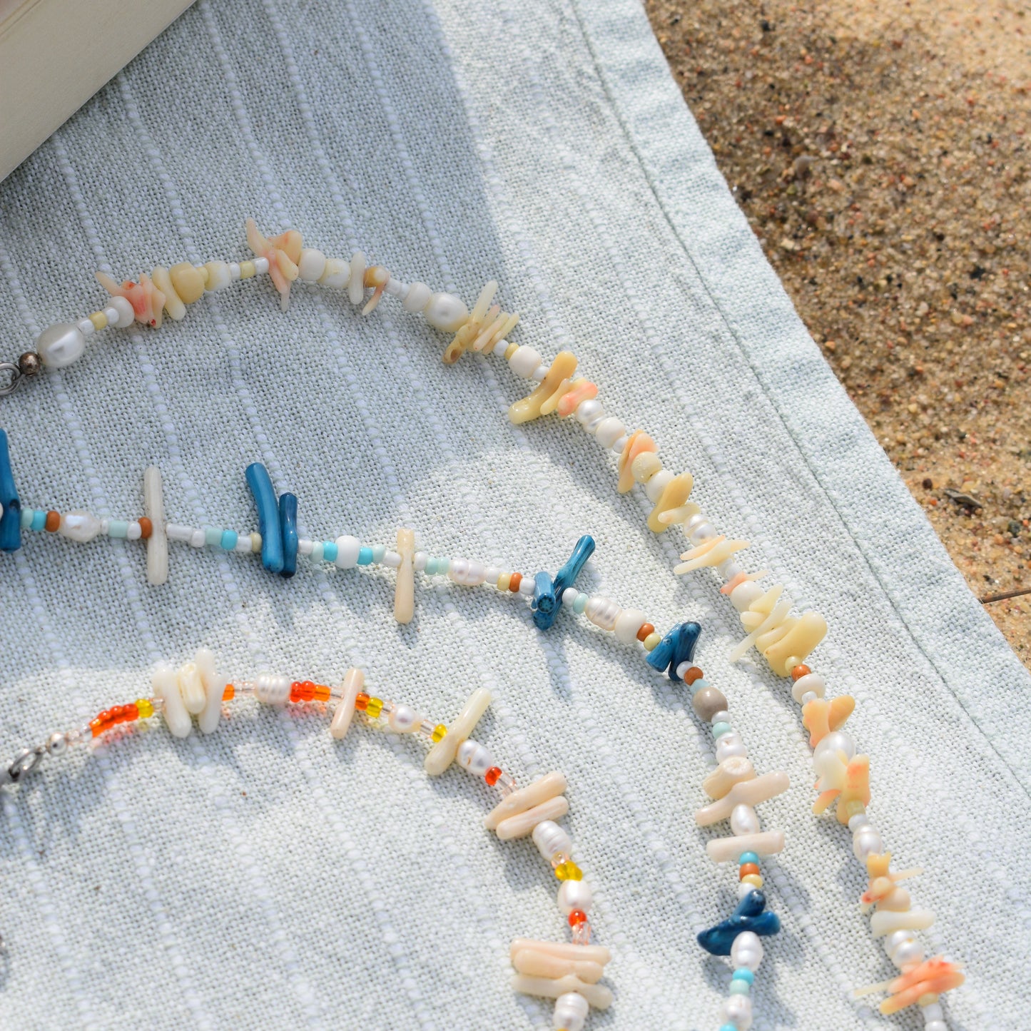 A close-up of Salty Dreamers Necklace | Ocean Bamboo Blue, a handmade piece with pearls, colorful glass beads, and shells, resting on light blue striped cloth beside sand.