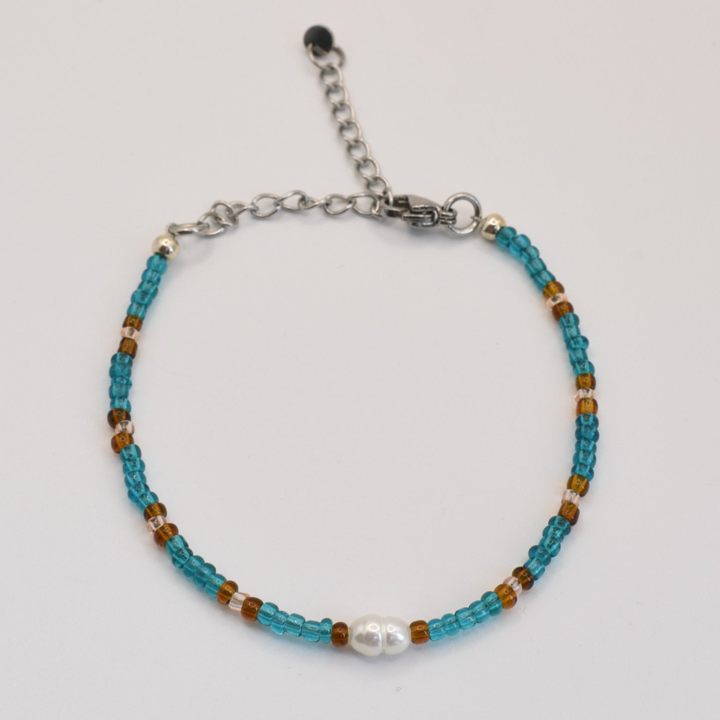 The Bracelet | Ocean Chique by Salty Dreamers features handmade turquoise and brown beads, a central white pearl-like bead, and an adjustable silver clasp—ideal for surfer chic style. Shown against a white background.