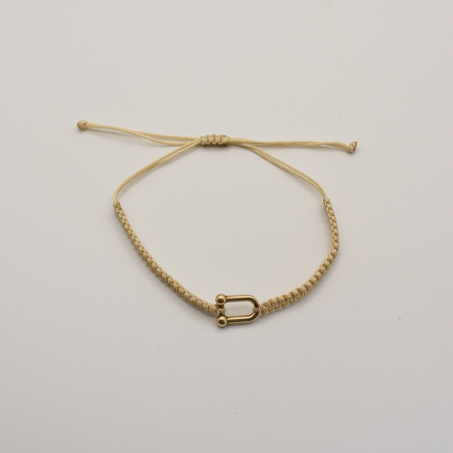 The Salty Dreamers Bracelet | Sailors Love Beige is a handmade beige macramé bracelet with a small gold-colored U-shaped charm, inspired by classic sailor shakel designs and shown on a plain white background.