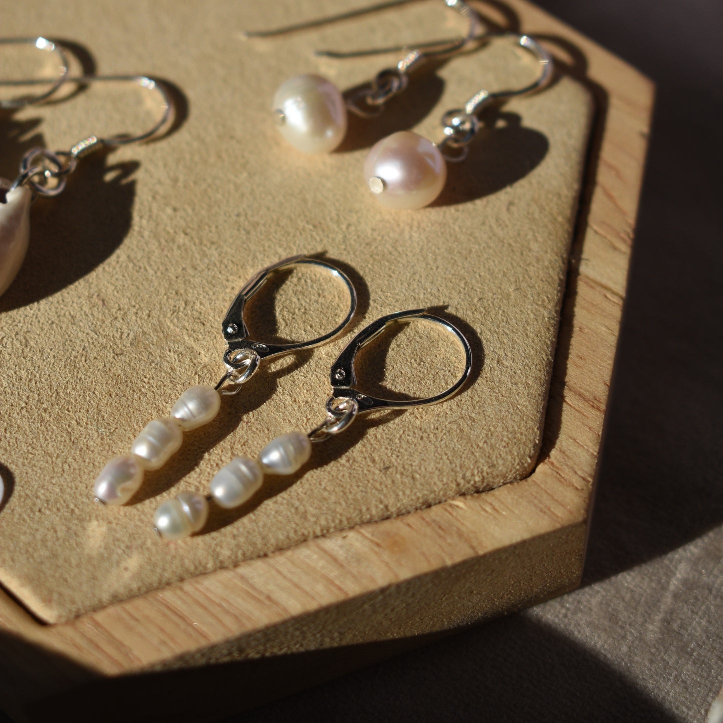 Two pairs of Salty Dreamers Pretty Pearly Earrings are displayed on a tan hexagonal tray. One pair has three freshwater pearls on 925 silver hoops, while the other features single round pearls on hooks, softly lit by sunlight.