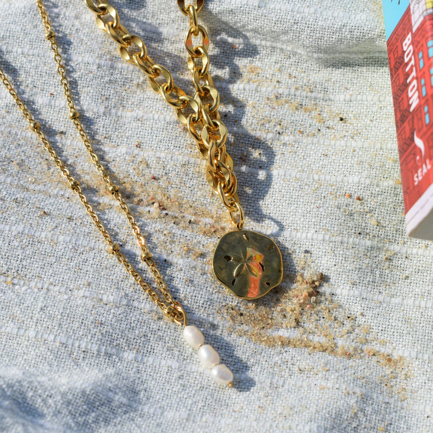 Two gold necklaces from Salty Dreamers rest on light textured fabric: one is the Ocean Dollar Gold necklace with a sand dollar pendant, and the other has a delicate chain with three small white pearls. Sand and part of a colorful book are nearby.
