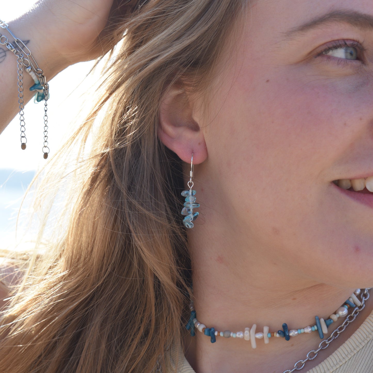 A woman with light brown hair smiles in the sunlight, wearing the Salty Dreamers Ocean Bamboo Blue necklace and matching blue and white beaded jewelry, including a bracelet and drop earrings, as she lifts her hand to her head.