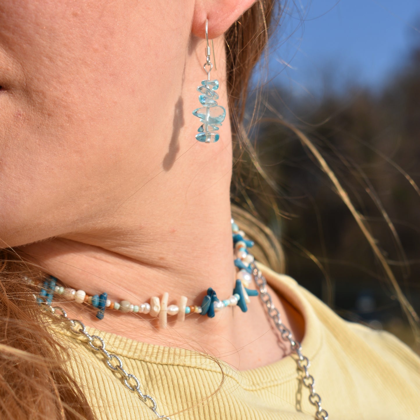 A close-up of a person wearing Salty Dreamers Necklace | Ocean Bamboo Blue, featuring blue stones and pearls, styled with a yellow shirt and sunlit hair.