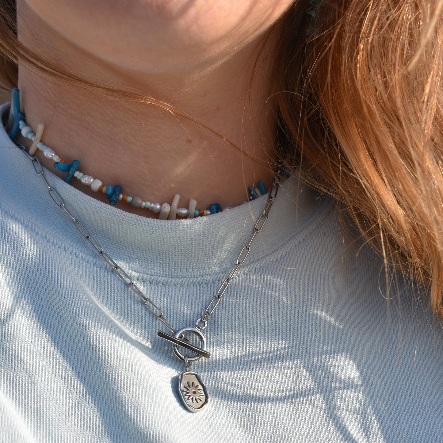 Close-up of a person wearing Salty Dreamers Necklace | Ocean Bamboo Blue—a blue and white glass bead choker—paired with two silver necklaces: one featuring a toggle clasp and the other an embossed pendant. Long, light brown hair partially visible.