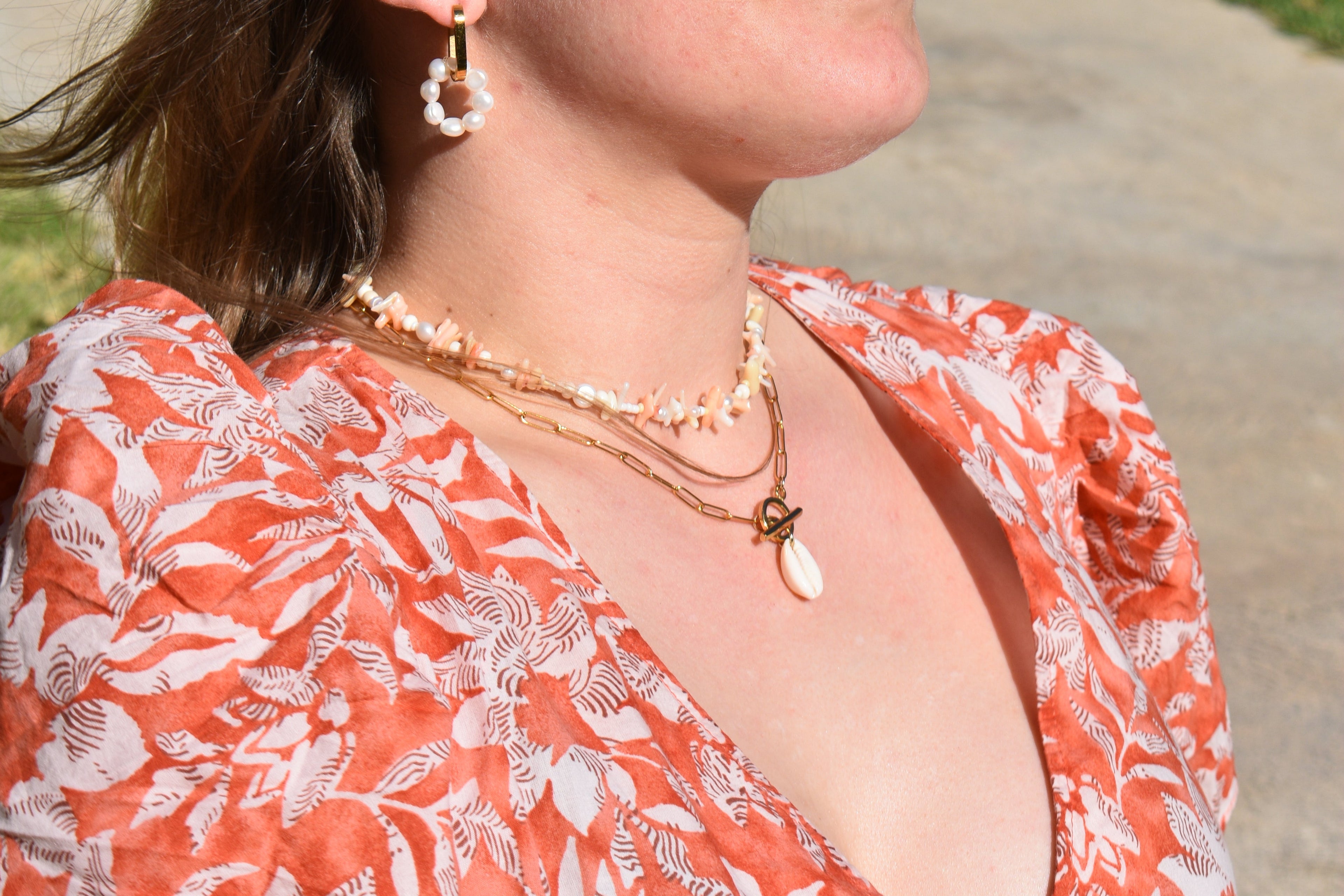 A woman wearing a red and white floral dress, layered seashell necklaces, and pearl hoop earrings stands outdoors in sunlight.