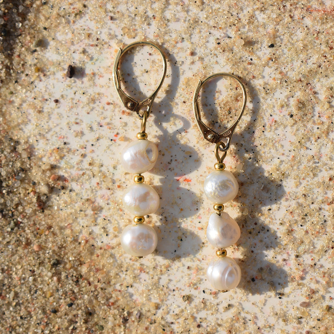 Salty Dreamers’ Earrings | Prettiest Pearly showcase three irregular freshwater pearls on each gold drop, photographed on sunlit sand—ocean-inspired jewelry capturing the unique beauty of handcrafted pearl earrings.