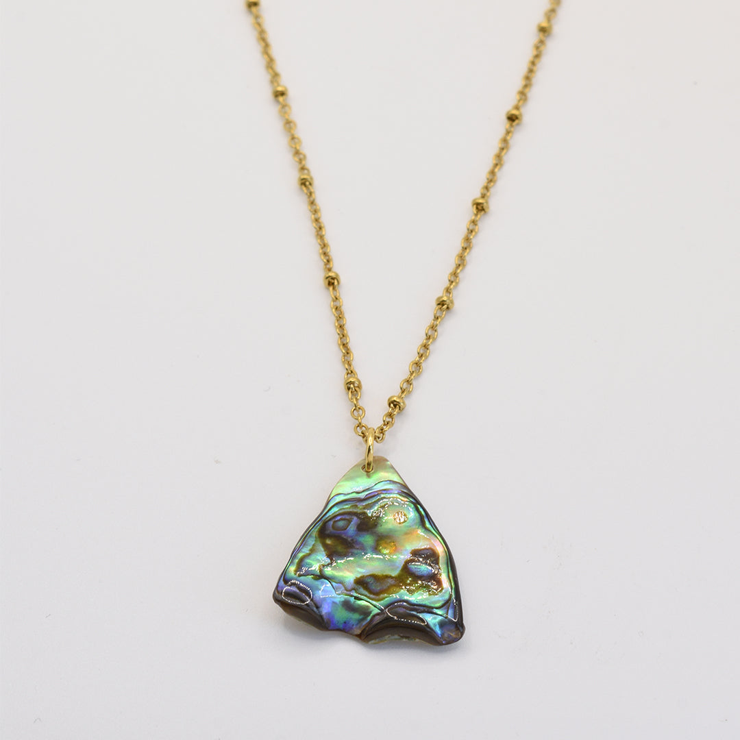 The Salty Dreamers Paua Shell Tai Necklace features a handmade pendant with an iridescent, irregular paua shell in green, blue, and purple hues, displayed on a gold chain against a plain white background.