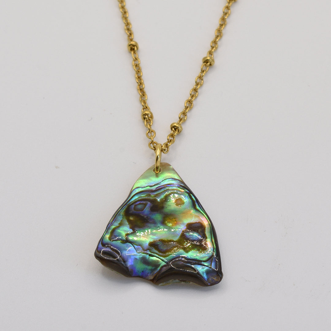The Salty Dreamers Necklace | Paua Shell Tai features a gold chain with a handmade pendant crafted from an irregular, iridescent pāua shell in green, blue, and purple hues. Shown against a white background.