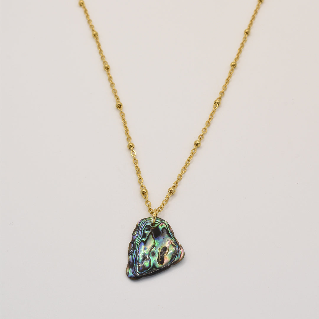 The Salty Dreamers Necklace | Paua Shell Moana features a gold chain with small beads and an iridescent triangular pāua shell pendant, inspired by New Zealand jewelry traditions. Shown on a plain white background.