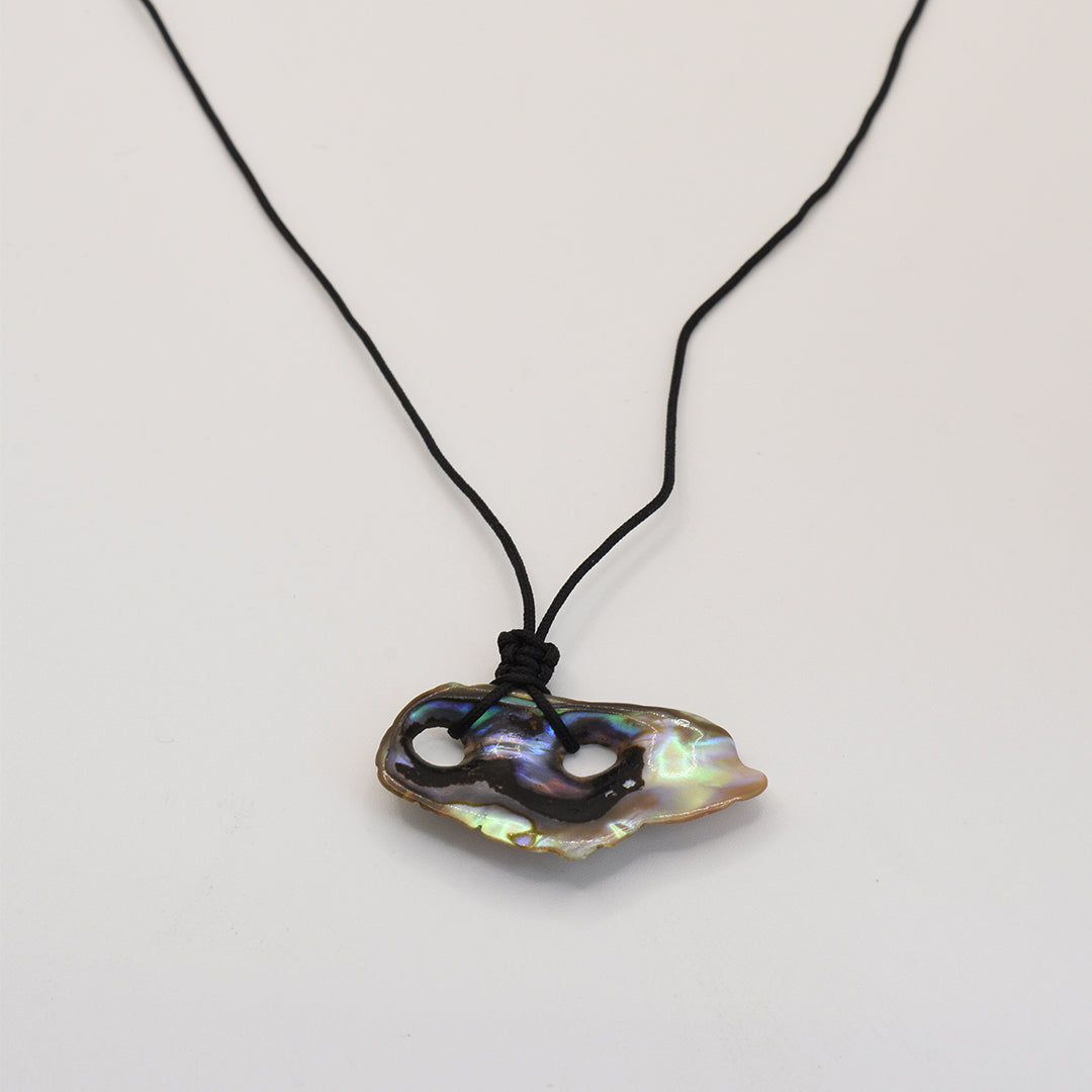 The Salty Dreamers Necklace | Paua Shell Haeata features a handmade black cord and an iridescent, irregularly shaped pāua shell pendant with two carved holes—an elegant New Zealand jewelry piece displayed on white.