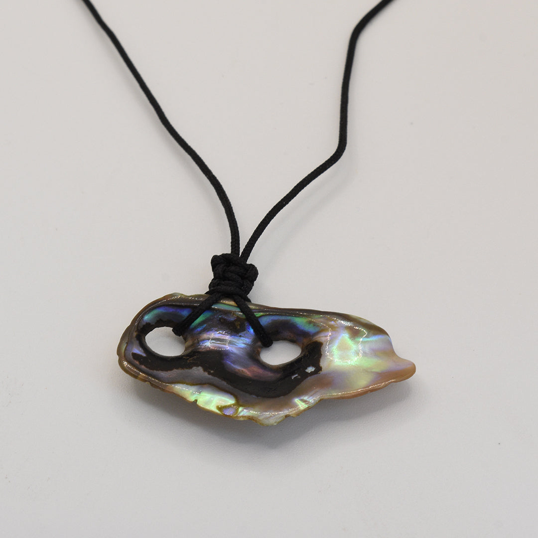 The Necklace | Paua Shell Haeata by Salty Dreamers features a handmade design with a black cord and an iridescent, irregular pāua shell pendant with two holes, shown on a plain white background.