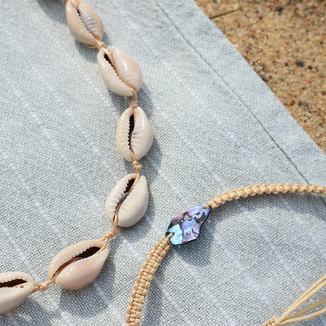 Close-up of the Salty Dreamers Cowrie Choker—a cowrie shell necklace resting on textured blue fabric by sandy ground. This necklace is perfect beach jewelry to elevate your summer style.