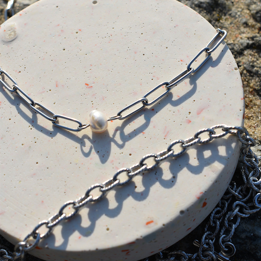 Two Salty Dreamers silver chain necklaces, including the handmade Paperclip Cutie Silver and a freshwater pearl design, are displayed on a round speckled stone surface with sunlight casting shadows and highlights on the chains.