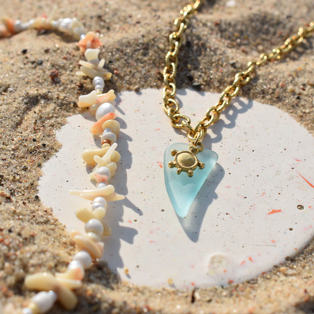 The Salty Dreamers Necklace | Ocean Bamboo Pink features a gold chain with a pink sea glass pendant and sun charm, styled on sand beside a seashell and handmade pearl beads, with sunlight highlighting its elegant details.