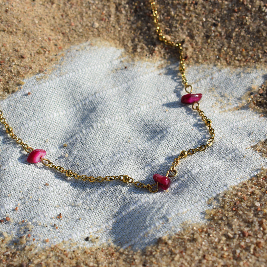 The Necklace | Shell Rubin by Salty Dreamers—a gold stainless steel chain with three small pink shell charms—rests on sandy fabric in bright sunlight, capturing ocean vibes.