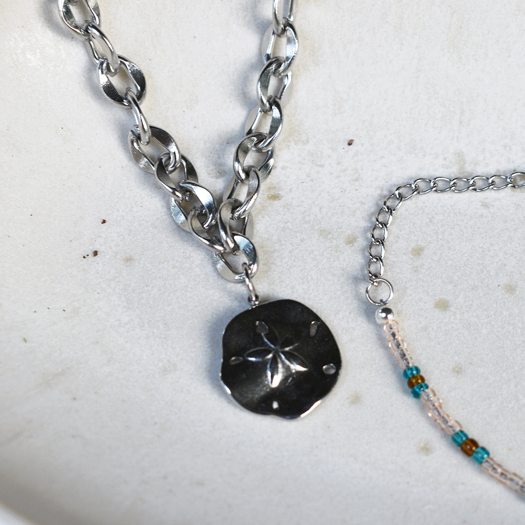 The Salty Dreamers Ocean Dollar Silver necklace with a chunky stainless steel chain and sand dollar pendant rests beside a delicate turquoise-accented beaded bracelet on a white ceramic dish.
