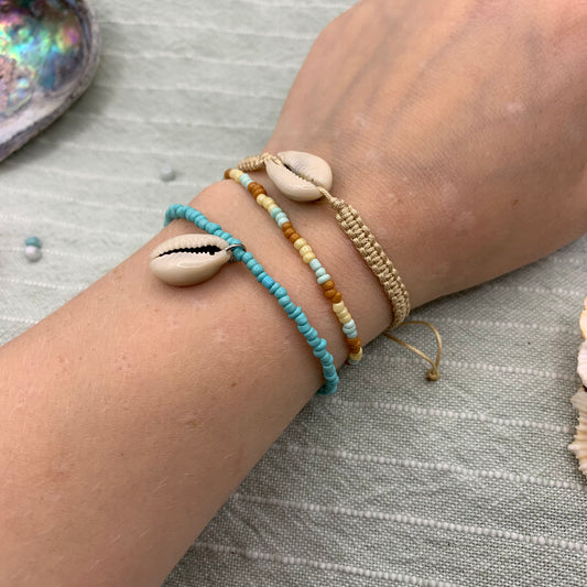 Hand trägt Salty Dreamers Armband Macramazing Cowrie Beige mit beigem Makramee-Band und weißer Kauri-Muschel.