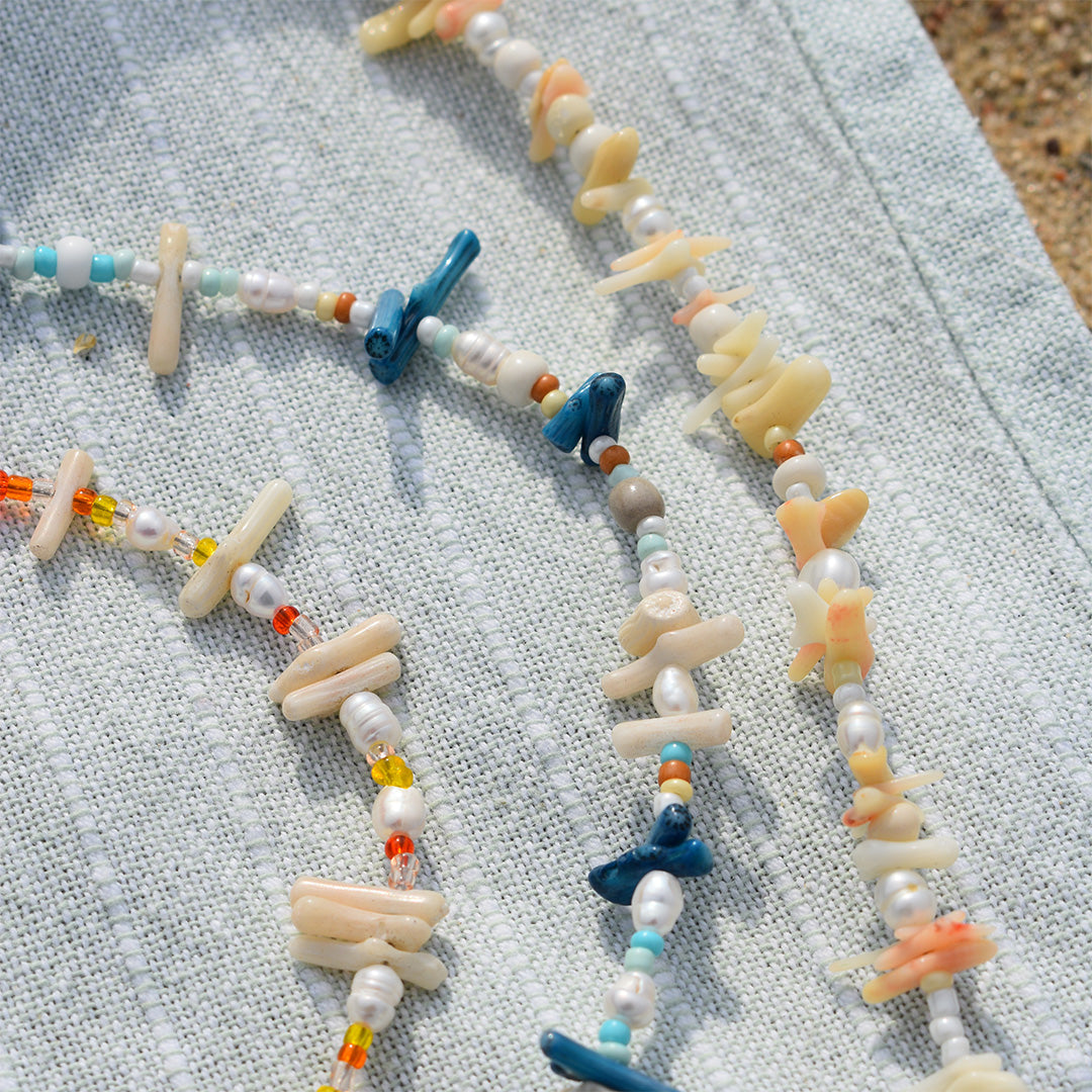 Beaded necklaces with white, blue, and orange beads and irregular shell pieces are displayed on a light blue textured fabric outdoors.