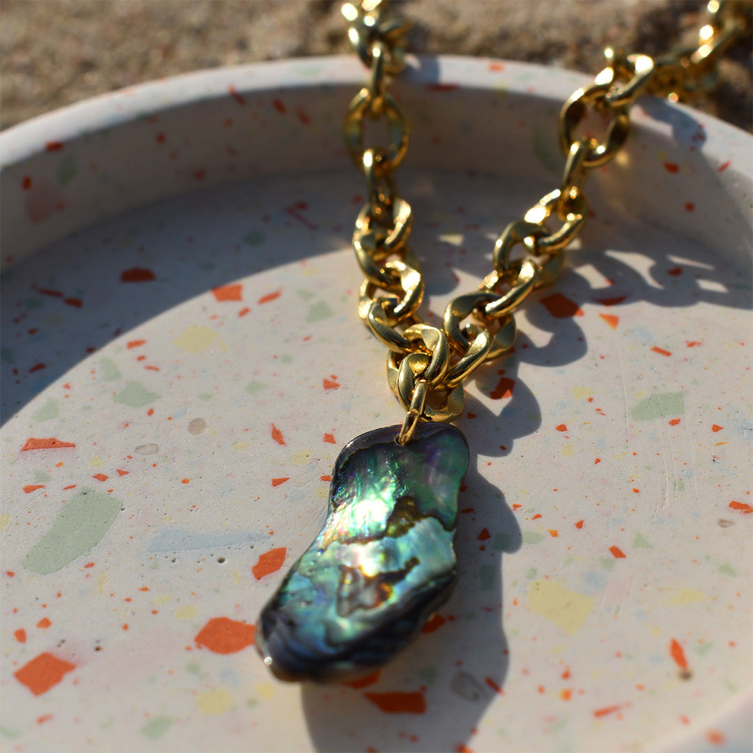 A chunky gold chain necklace with an iridescent abalone shell pendant rests on a speckled terrazzo dish in sunlight.