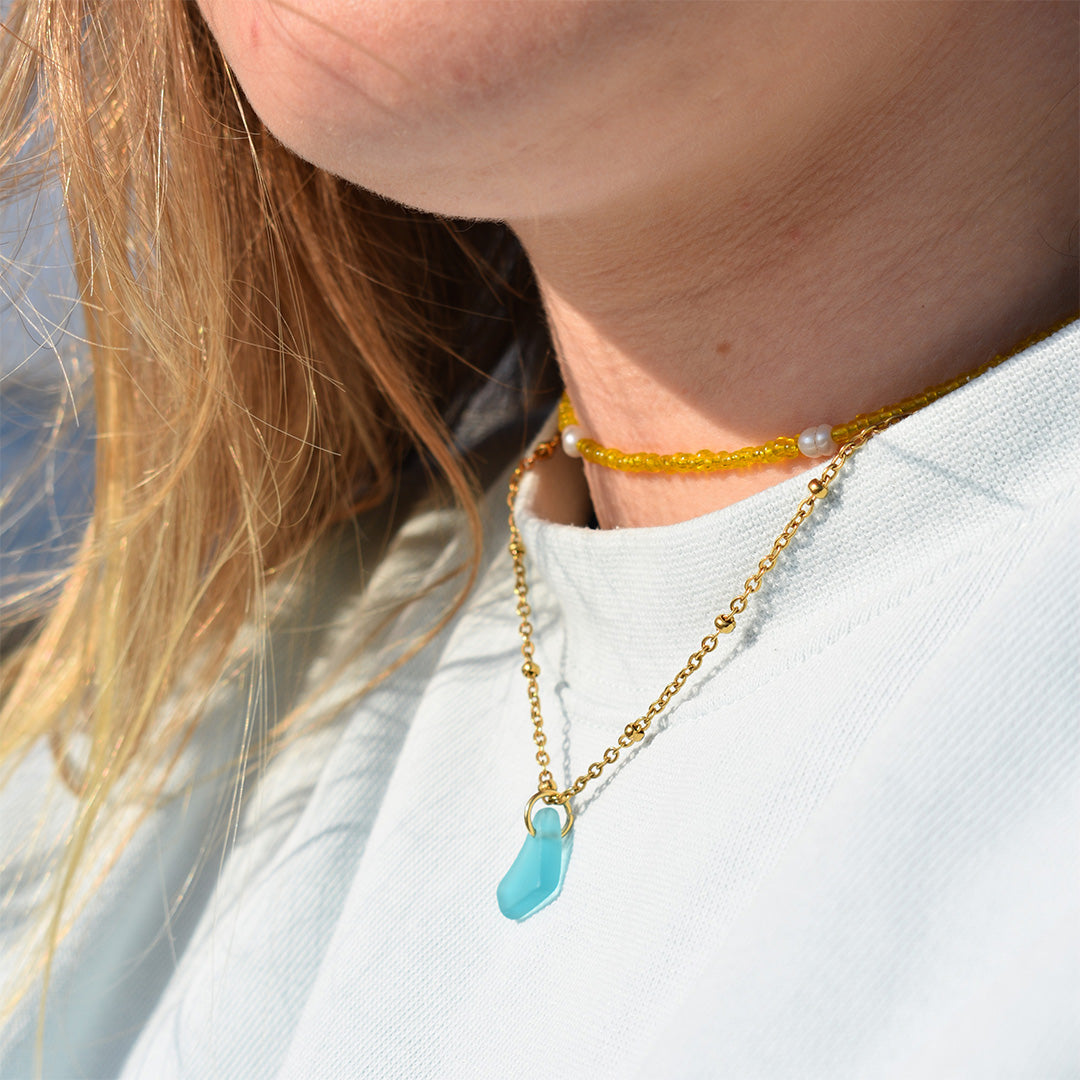 A person wearing two necklaces: one is a yellow beaded choker with a small pearl, and the other is a gold chain with a blue geometric pendant. The person is dressed in a light-colored top.
