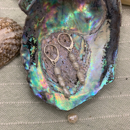 Pretty Pearly earrings by Salty Dreamers feature freshwater pearls and 925 silver hooks, presented in a colorful abalone shell on textured beige fabric with seashells nearby.