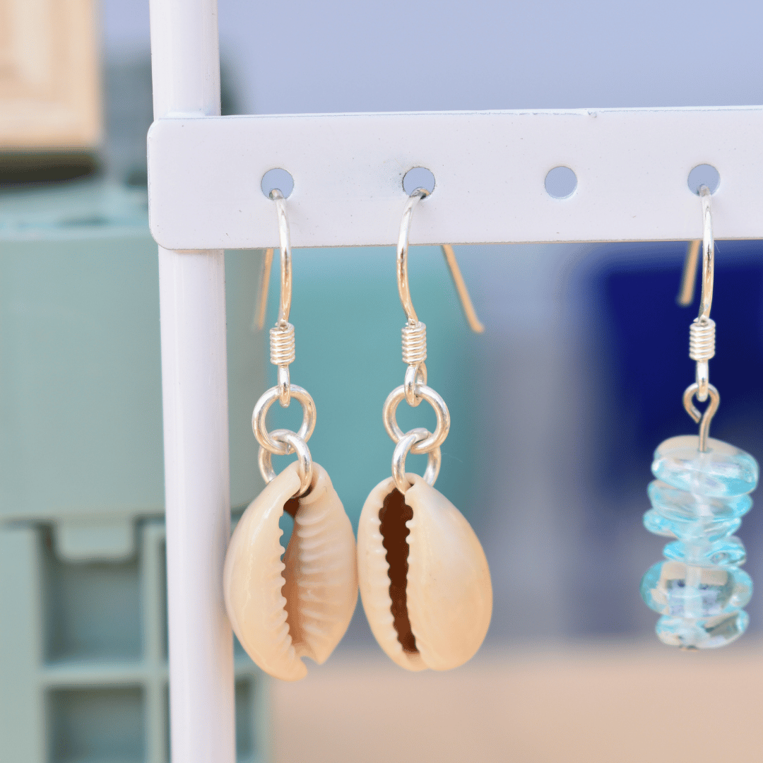 A pair of Salty Dreamers Dangling Cowrie Silver earrings with beige shells hang on a white rack beside another handmade pair featuring stacked blue glass beads.