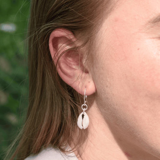 A close-up shows a person wearing Salty Dreamers' Earrings | Dangling Cowrie Silver—925 silver hoops with white cowrie shell pendants—standing out against a softly blurred green background.