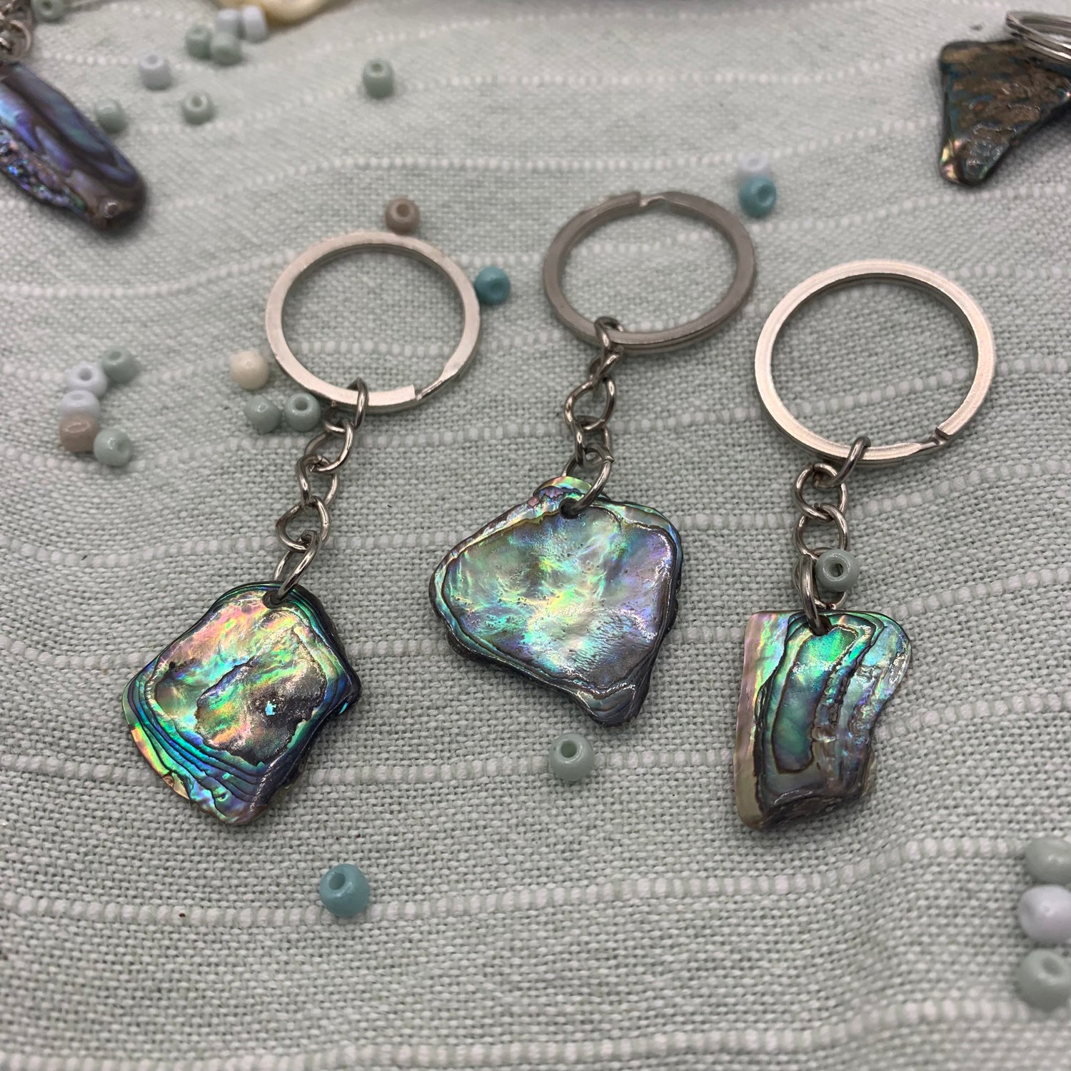 Three Salty Dreamers Keychain | Paua Shell keychains are displayed on light woven fabric, surrounded by pastel beads and shell fragments.