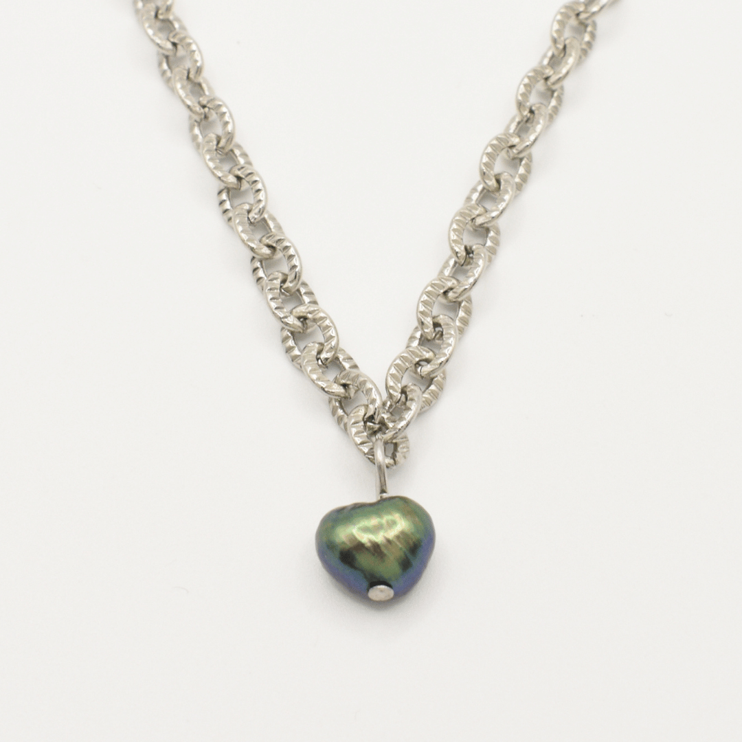 The "A Beach Pearl" necklace by Salty Dreamers features a silver chain and a dark iridescent heart pendant with a subtle surfer vibe, showcased against a white background.