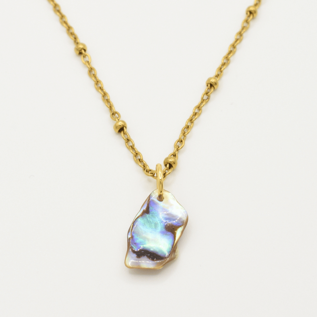 The Salty Dreamers Necklace | Paua Shell Ra features a gold chain with small round beads and an irregular iridescent pāua shell pendant—handmade jewelry shown on a plain white background.