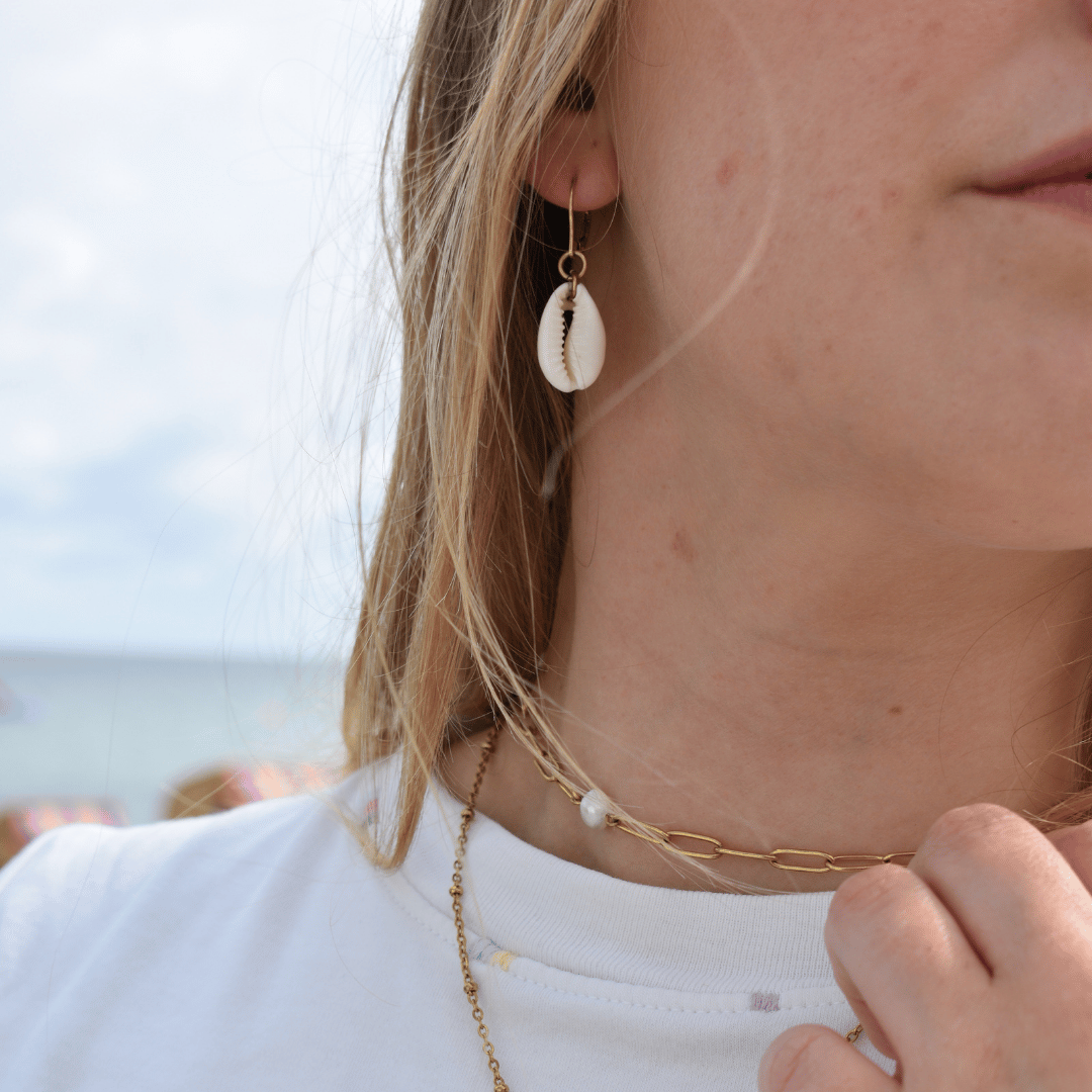 A woman wearing Salty Dreamers' Dangling Cowrie Gold earrings and a gold-plated 925-silver chain necklace with a white bead holds her necklace. The blurred background shows a beach and ocean scene.