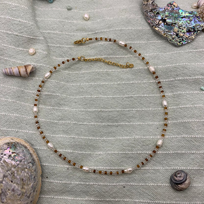 A handmade necklace with a combination of glass beads and freshwater pearls, featuring a golden stainless steel chain, displayed against a textured green background with seashells and other small items.