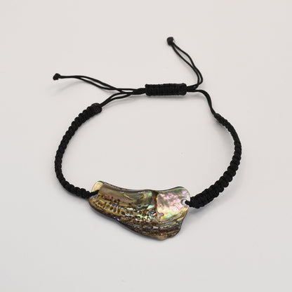 The Salty Dreamers Bracelet | Paua Shell Ngakau is a black braided handmade piece with an iridescent, irregular New Zealand abalone shell centerpiece, shown on a plain white background.