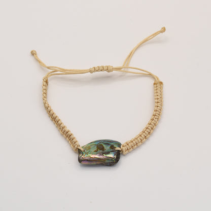 The Bracelet | Paua Shell Rangi by Salty Dreamers is a handmade beige macrame bracelet with a polished, irregular pāua shell bead at the center, shown on a plain white background.