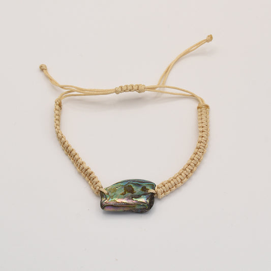 The Bracelet | Paua Shell Rangi by Salty Dreamers is a handmade beige macrame bracelet with a polished, irregular pāua shell bead at the center, shown on a plain white background.