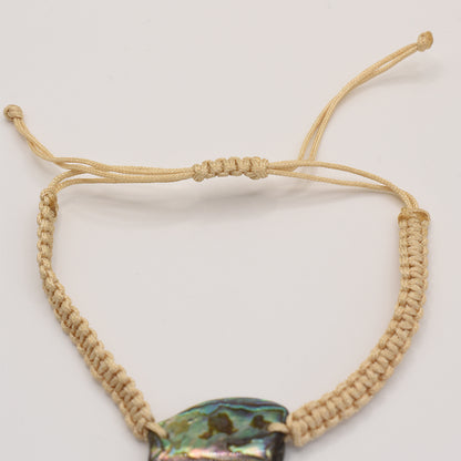 A close-up of the Salty Dreamers Bracelet | Paua Shell Rangi, featuring a handmade beige macrame band and a vibrant iridescent pāua shell centerpiece against a plain white background.