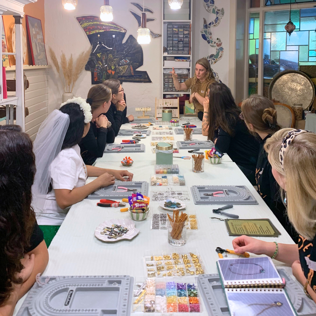 A group of women gather around a table filled with jewelry-making supplies, listening to an instructor at the Workshop Meeresschmuck in Kiel by Salty Dreamers in a brightly decorated craft studio.
